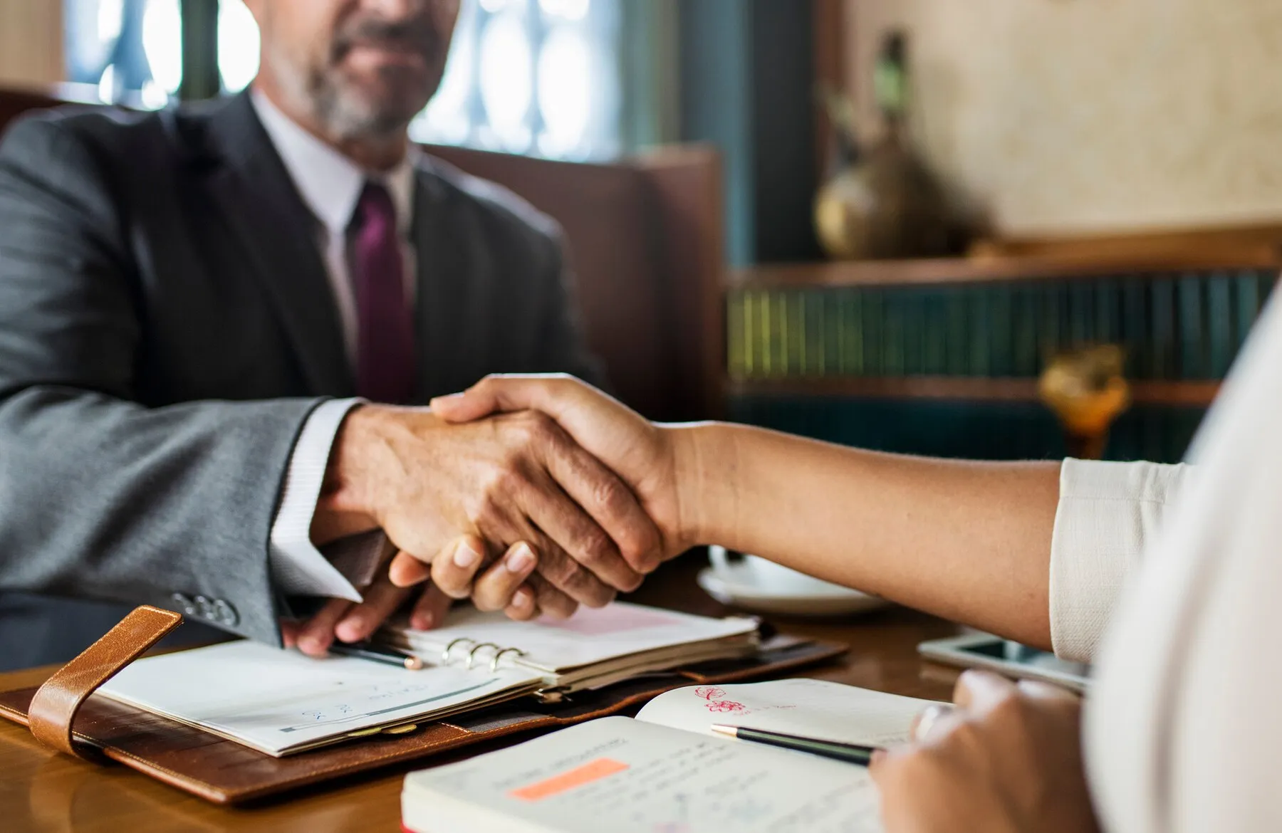 Man in suit shaking hands with individual with a desk and paperwork below.