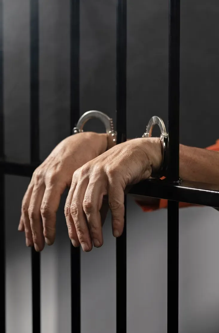 Individual in jail cell with handcuffs on stick them on of the cell.
